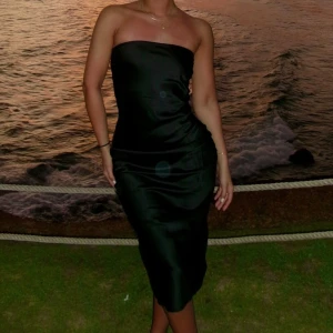 Svart axelbandslös satinklänning - Säljer en svart axelbandslös klänning i satin med snygg slits bak och knytning i ryggen. Klänningen har en tight passform och elegant glansigt material som ger en lyxig känsla. Perfekt för dig som vill sticka ut med en stilren look.