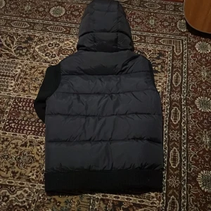 Svart dunväst med huva från Moncler - Snygg svart dunväst från Moncler med stickade sidor och ärmar, quiltad framsida och baksida samt stor vadderad huva. Västen har dragkedja framtill och Moncler-logga på ärmen. Perfekt för lager-på-lager och streetstyle.
