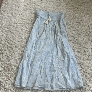 Ljusblå långklänning från Gina Tricot - Supersöt ljusblå långklänning från Gina Tricot med smock upptill och knytband med tofsar. Klänningen har ett diskret mönster och är perfekt för varma dagar. Luftigt material och snygg passform som ger en chill vibe.