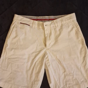 Beiga shorts från Tommy Hilfiger - Snygga beiga shorts från Tommy Hilfiger i ekologisk bomull. Klassisk modell med bälteshällor, fickor fram och bak samt diskret logga ovanför ena bakfickan. Perfekta för varma dagar och enkel att matcha med det mesta.