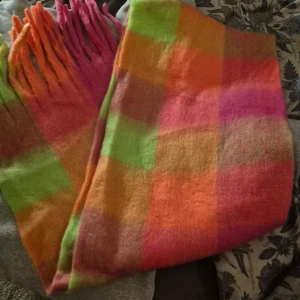 Färgglad rutig halsduk med fransar - En stor och fluffig halsduk med rutigt mönster i neonrosa, orange, grön och gul. Halsduken har långa fransar i matchande färger och är perfekt för att liva upp vinterlooken. Materialet känns mjukt och värmande, perfekt att svepa in sig i kalla dagar.