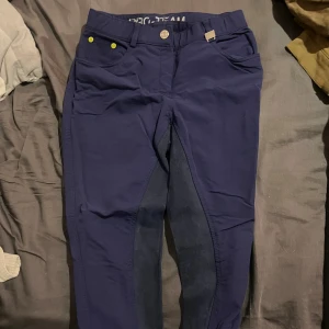 Mörkblå ridbyxor från Pro Team - Snygga mörkblå ridbyxor från Pro Team med förstärkta partier på insidan av benen för extra grepp. Byxorna har flera fickor, detaljer med knappar och en bekväm midja. Perfekta för dig som gillar ridning och vill ha stil på stallet.