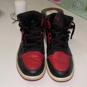 Nike Air Jordan 1 Bred Svart/Röd - Säljer ett par ikoniska Nike Air Jordan 1 i färgen Bred med svart och rött skinn, klassisk vinge-logga på sidan och svart snörning. Mellanhöga sneakers med vit sula och röd yttersula. Perfekta för dig som gillar streetstyle och vill sticka ut.