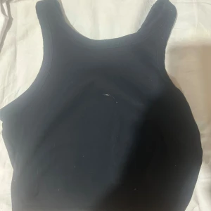 Svart ribbad linnetopp från 157 - En svart ribbad linnetopp från 157 med klassisk rak halsringning och breda axelband. Toppen är ärmlös och har en enkel, stilren design som passar perfekt till jeans eller kjol. Materialet är mjukt och stretchigt för en bekväm passform.