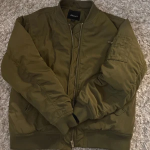 Olivgrön bomberjacka från Monki - Säljer en olivgrön bomberjacka från Monki med klassisk passform. Jackan har dragkedja framtill, ribbade muddar och en ärmficka med dragkedja. Perfekt för dig som vill ha en stilren och trendig jacka till vardags.