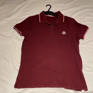 Vinröd pikétröja från Moncler S - Vinröd pikétröja från Moncler i klassisk modell med vit och röd rand på krage och ärmslut. Tröjan har broderad Moncler-logga på bröstet och knäppning med tre knappar. Tillverkad i bomull och passar perfekt till en snygg och avslappnad stil.