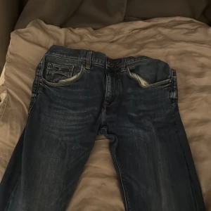 Tommy Hilfiger jeans blå slim fit - Säljer ett par snygga Tommy hilfiger jeans i mörkblå tvätt med slitningar och kontrastsömmar. Modellen är slim fit med klassiska fem fickor och detaljerade nitar. Jeansen har orange och mörkblå sömmar på insidan av benslutet.