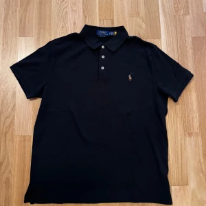 Svart pikétröja från Polo Ralph Lauren - Klassisk svart pikétröja från Polo Ralph Lauren med korta ärmar och krage. Tröjan har tre vita knappar framtill och den ikoniska broderade loggan på bröstet. Perfekt för en clean och stilren look.Som ny