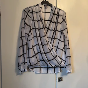 Vit omlottblus med svart rutmönster H&M - Snygg vit omlottblus från H&M i storlek 38 med svart rutigt mönster. Blusen har lång ärm, v-ringad omlottfront och en liten knapp på insidan för att hålla omlotten på plats. Materialet är lätt och luftigt, perfekt för en cool och modern look.