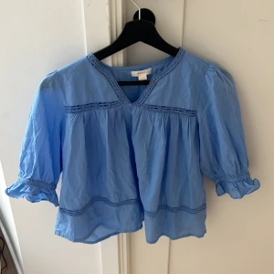 Blå blus med spets från Gina Tricot - Supersöt ljusblå blus från Gina Tricot med v-ringning och spetsdetaljer vid halsen och över bröstet. Blusen har puffiga korta ärmar med volangkant och är tillverkad i mjuk bomull. Perfekt för dig som gillar romantisk och chill stil.