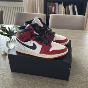 Nike Air Jordan 1 Chicago Mid - Nu säljer jag mina riktigt feta Nike Air Jordan 1 Chicago Mid med klassisk färgblockning i rött, vitt och svart. Skorna har svart swoosh, perforerad tå, högt skaft och snörning. Tillverkade i skinn. Levereras med originalkartong och extra svarta skosnören. Skorna är som nya. 