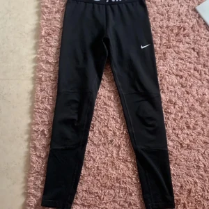 Nike Pro svarta leggings strl L - Svarta Nike Pro leggings med vit logga på benet och bred resår med Nike-tryck i midjan. Snygg, tight passform som sitter skönt och är perfekt för träning. Materialet är stretchigt och andas, vilket gör dem grymma för gymmet eller sport.