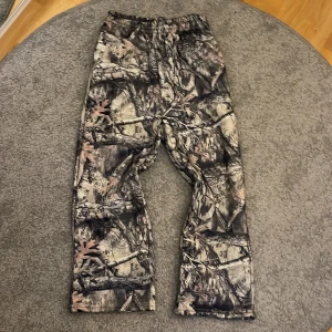 Camouflage täckbyxor från Akimbo - Säljer ett par feta täckbyxor från Akimbo med Realtree-camomönster. Byxorna har flera praktiska fickor och elastisk midja för skön passform. Perfekta för dig som vill sticka ut med coolt camo och vara redo för utomhusäventyr.