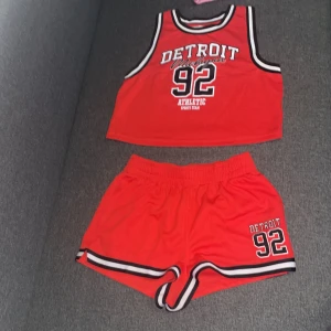 Röda sportiga shorts+linne Detroit 92 - Snygga röda shorts+linne med svart och vit rand längs kanten, elastisk midja och tryck 'Detroit 92' på benet. Perfekt för dig som gillar sportig stil och vill sticka ut med en cool basketinspirerad look. Matcha med tillhörande linne för en komplett outfit.