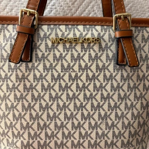 Michael Kors beige handväska med logomönster - Snygg handväska från Michael Kors i beige och grått med MK-logga över hela väskan. Väskan har bruna handtag med guldfärgade spännen och detaljer. Medföljande axelband💞