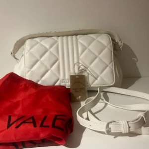 Snygg (Mario) Valentino handväska - Fin vit (Mario) Valentino handväska modell Soda Bianco. Material: polyester Huvudstängning: magnetisk Handtag: 1 handtag Insida: 3 fodrade fack Innerfickor: 1 Bredd cm: 30 Höjd cm: 17 Djup cm: 12 Köpt våren 2022 och anvönd vid ett tillfälle. Mycket fint skick förutom lite slitage undertill som man inte tänker på. Se bild 5. Dustybag medföljer. Tag och kvitto finns att få med om så önskas. Inköpspris: 1068 kr. Hämtas i Göteborg eller Kungälv.  Skickas om köparen betalar för frakten.