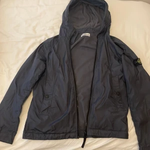 Mörkblå vindjacka från Stone Island - Snygg mörkblå vindjacka från Stone Island med huva och klassisk patch på vänster ärm. Jackan har två fickor med knappstängning och dragkedja framtill. Perfekt för dig som gillar streetwear och vill ha en clean look. Priset går att diskutera över 