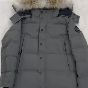 Grå dunjacka från Canada Goose Wyndham - Säljer en grå dunjacka från Canada Goose med fluffig päls på huvan och svart logotyp på ärmen. Jackan har långa ärmar, dragkedja och knappar framtill, samt två sidofickor med dragkedja. Perfekt för kalla vinterdagar och riktigt snygg stil. (Helt ny)