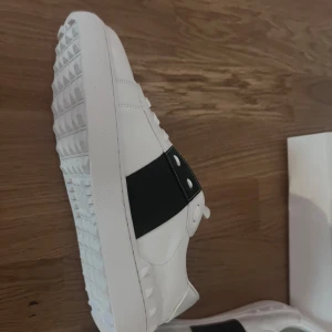 Valentino Garavani Open sneakers vit/svart - Säljer ett par Valentino Garavani Open sneakers i vitt skinn med bred svart läderdetalj över vristen. Snygg chunky sula med pyramidmönster och diskret logga på plösen. Perfekta för dig som vill ha en clean men lyxig look.