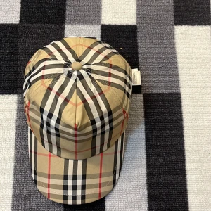 Burberry rutig keps i beige och svart ( pris kan diskuteras ) - Snygg keps från Burberry med klassiskt rutigt mönster i beige, svart, vitt och röda linjer. Kepsen har böjd skärm och justerbar rem baktill. Tillverkad i bomull och polyester, perfekt för att lyfta din streetwear-look.