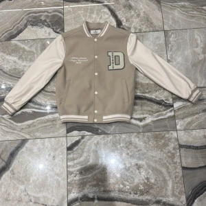 Beige och vit bomberjacka Detroit - Snygg beige och vit bomberjacka från Detroit med broderad text 'Athletic Division League Tournament 1993' och stort D-märke på bröstet. Jackan har långa ärmar, ribbade muddar och knappar framtill. Perfekt för dig som gillar retro college-vibe.
