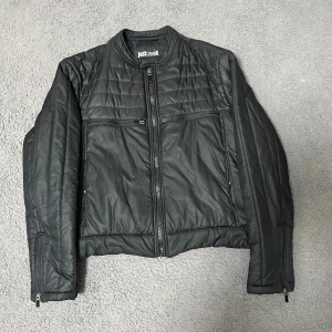 Svart quiltad pufferjacka Just Cavalli - Svart pufferjacka från Just Cavalli med quiltade paneler och broderad logga på ryggen. Jackan har flera dragkedjor framtill, ståkrage och långärmade ärmar med dragkedja vid ärmslut. Snygg och modern look som passar till streetwear.