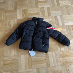 Svart Moncler pufferjacka med ränder - Svart pufferjacka från Moncler med klassisk quiltad design och dragkedja framtill. Jackan har långa ärmar och en snygg detalj med röd, vit och svart rand på ena ärmen samt Moncler-logga. Perfekt för kalla dagar och riktigt stilren look.