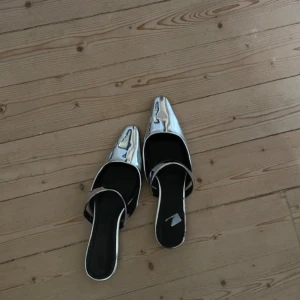Silvriga slip-in ballerinaskor - Säljer ett par riktigt snygga ballerinaskor i metallic silver med spetsig tå och platt sula. Skorna har öppen häl och en bred rem över foten. Perfekta för dig som vill sticka ut med en futuristisk vibe och gillar att mixa material och former.