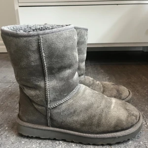 Grå UGG boots i mocka - Mysiga grå UGG boots med rund tå och platt sula. Utsidan är i mjuk mocka och insidan är fodrad med fluffigt syntetmaterial som håller fötterna varma. Klassisk UGG-logga baktill och grov yttersula för extra komfort. Perfekt för kalla dagar.