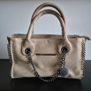 Beige handväska Stella McCartney - Snygg beige handväska från Stella McCartney med kedjedetaljer i silver och dekorativa sömmar längs kanterna. Väskan har dubbla handtag och är tillverkad i syntetmaterial med en stilren, boxig form. Perfekt för dig som gillar trendiga accessoarer med edge.