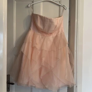 Ljusrosa volangklänning Coast - Superfin ljusrosa kortklänning från Coast med flera lager volanger och axelbandslös design. Klänningen har en romantisk look med mjukt skimrande tyg och vackra draperingar upptill. Perfekt för dig som vill sticka ut med en feminin och fluffig stil.