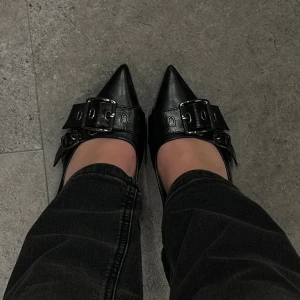 Svarta pumps med spänne från Claudia Ghizzani - Säljer ett par svarta pumps från Claudia Ghizzani med spetsig tå och stora metallspännen. Skorna har en platt sula och är tillverkade i skinnimitation. Snygg och edgy design som sticker ut med sina detaljer.