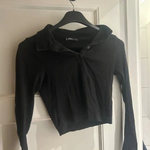 Svart croppad ribbad topp FB Sister - Säljer en svart croppad topp från FB Sister i storlek M. Toppen har lång ärm, krage och knappar framtill. Materialet är ribbad bomull som ger en skön och snygg känsla. Perfekt till jeans eller kjol för en chill och trendig look.