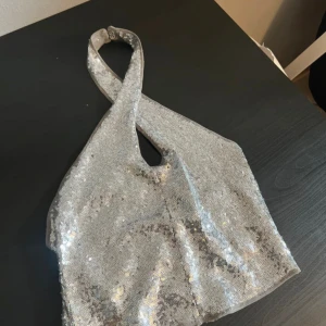 Silvrig glittrig axelväska - Snygg silvrig axelväska med glittriga paljetter över hela ytan. Väskan har en unik form med korsad axelrem och ett litet hål framtill som detalj. Perfekt för att lyfta din outfit med extra bling och cool vibe.