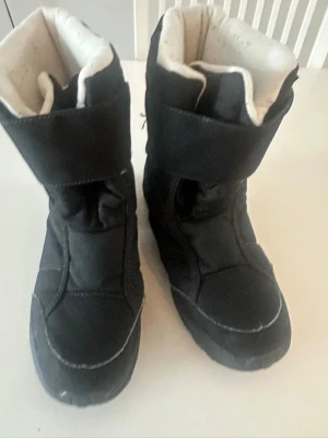 Svarta vinterstövlar  - Svarta vinterstövlar från Quechua med kardborreband och vadderad insida i vitt. Skorna är vattentäta och har en robust sula för bra grepp. Tillverkade i syntetmaterial och designade för att hålla fötterna varma under kalla dagar.