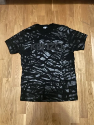 Svart zebramönstrad Kenzo t-shirt - Säljer en svart t-shirt från Kenzo med blankt zebramönster och stor logga över bröstet. Klassisk passform med korta ärmar och rund hals. Materialet känns mjukt och skönt, perfekt för dig som vill sticka ut lite extra.