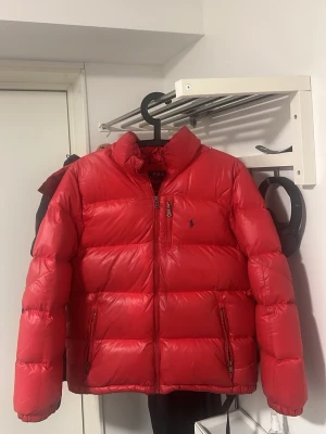 Röd Ralph Lauren dunjacka – stl 14–16 - Röd Ralph Lauren dunjacka – stl 14–16  Säljer en röd, glansig Ralph Lauren dunjacka i storlek 14–16. Klassisk modell med luva och broderad logga på bröstet.  ✔️ Varm och skön ✔️ Glansigt material ✔️ Luva ✔️ Original Ralph Lauren  Skick: – Två små hål som är lagade – Klädhängaren är sönder men mycket lätt att laga – I övrigt bra skick  Pris kan diskuteras vid snabb affär. Skriv vid intresse