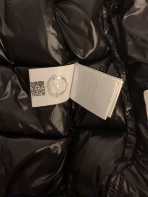 Svart Moncler dunväst med huva - Säljer en svart, glansig dunväst från Moncler med huva och två dragkedjeförsedda fickor. Västen har Moncler-logga på bröstet och coolt serietryck på insidan. Perfekt för lager-på-lager och riktigt snygg till streetwear-stilen.