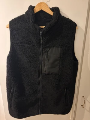Svart fleeceväst från True North - Mysig svart fleeceväst från True North med hög krage och hel dragkedja. Västen har en bröstficka med dragkedja i nylonmaterial och två sidofickor med dragkedja. Perfekt för lager-på-lager och chill dagar. Skriv för mer info! ABS! Pris går att diskutera.