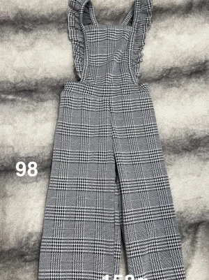 Rutiga byxdress med volangdetaljer - Säljer en snygg byxdress i svartvitt rutigt mönster. Byxdressen har breda axelband med volangdetaljer och raka, vida ben. Perfekt för dig som gillar trendiga och bekväma plagg med lite extra detaljer.🌸Använda två gånger. Storlek 98