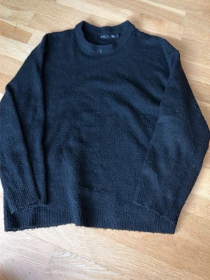 Svart stickad tröja med rund hals - En klassisk svart stickad tröja med rund hals och långa ärmar. Tröjan har ribbade muddar vid ärmslut och nederkant, vilket ger en enkel och stilren look. Perfekt att slänga på sig när det är kyligt ute.