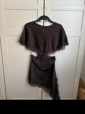 Brun asymmetrisk klänning med spets - Supersnygg svart kortklänning från H&M med asymmetrisk nederkant och spetsdetaljer längs kanterna. Klänningen har cut-out i midjan och cape-liknande överdel som ger en unik look. Perfekt för dig som vill sticka ut med en trendig och elegant stil.