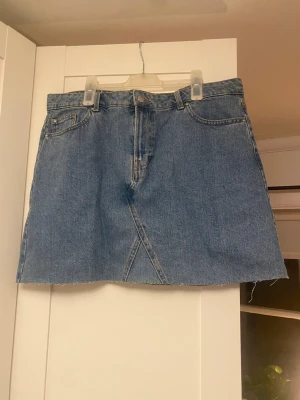 Blå jeanskjol från Denim Co. - Säljer en klassisk blå jeanskjol från Denim Co. med rå kant nertill och femficksmodell. Kjolen har en snygg sömdetalj framtill och är i en rak, kort modell. Perfekt till sommaren!