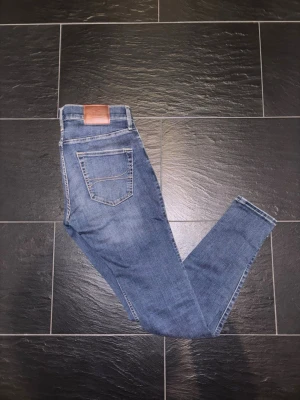 Blå slitna jeans från Hollister W31 L32 - Säljer ett par blå jeans från Hollister med snygga slitningar och tvättade detaljer. Klassisk femficksmodell med normal passform och raka ben. Jeansen har kontrastsömmar och patch i läderimitation bak i midjan. Perfekta för en avslappnad look.