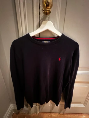 Mörkblå tröja från Polo Ralph Lauren - Klassisk mörkblå tröja från Polo Ralph Lauren i mjuk pima bomull. Tröjan har rund halsringning med röd rand på insidan och den ikoniska röda logon broderad på bröstet. Perfekt till jeans eller chinos för en clean och stilren look.