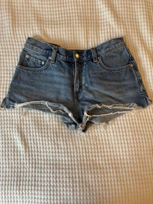 Lågmidjade jeansshorts  - Säljer ett par klassiska blå denimshorts med låg midja och råa, fransiga kanter. I ”jeanstyg”. Helt nya men endast testade + tvättade.