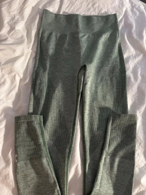Grågröna seamless leggings - Säljer ett par grågröna seamless leggings med hög midja och ribbade detaljer längs sidorna och bensluten. Materialet är stretchigt och mjukt, perfekt för träning eller chill. Diskret logga på ena vaden. Snygg passform som sitter tajt.