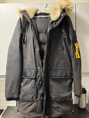 Svart parkajacka från Diesel med päls  - Säljer en svart parkajacka från Diesel som är äkta!  med stor huva och päls i gult och grått. Jackan har flera stora fickor, dragkedja och knappar framtill samt en gul detalj på ärmen. Insidan av huvan är fodrad med teddy. Perfekt för kalla dagar.