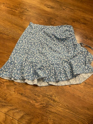 Blommig blå omlottkjol  - Supersöt blå kortkjol från SHEIN med vitt blommönster. Kjolen har omlottdesign, volangkant och knytband i sidan för extra detalj. Har även dragkedja på sidan. 