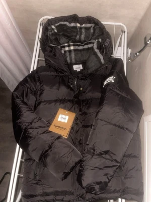 Svart pufferjacka från Burberry - Säljer en svart pufferjacka från Burberry med klassiskt rutigt foder i huvan. Jackan har dragkedja, knappar och två fickor framtill. Stort Burberry-tryck på ärmen och en riktigt clean look. Perfekt för kalla dagar och riktigt snygg till streetwear.